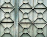 A metal gate featuring a geometric design with repeating patterns of circles and diamonds. The surface appears weathered and slightly rusty, with a faded bluish-green color.