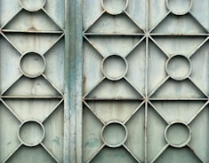 A metal gate featuring a geometric design with repeating patterns of circles and diamonds. The surface appears weathered and slightly rusty, with a faded bluish-green color.