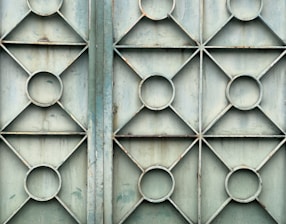 A metal gate featuring a geometric design with repeating patterns of circles and diamonds. The surface appears weathered and slightly rusty, with a faded bluish-green color.