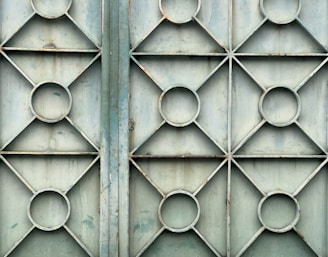 A metal gate featuring a geometric design with repeating patterns of circles and diamonds. The surface appears weathered and slightly rusty, with a faded bluish-green color.