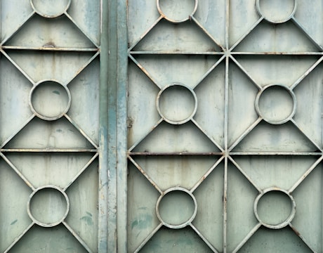 A metal gate featuring a geometric design with repeating patterns of circles and diamonds. The surface appears weathered and slightly rusty, with a faded bluish-green color.
