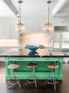 A modern kitchen with a central island painted in a bright green color, topped with a black marble countertop. Three wooden bar stools with metal legs are placed in front of the island. Above the island hang two spherical pendant lights with visible filament bulbs. A decorative blue bowl with intricate patterns sits on the countertop, and a stack of books is placed next to it. The kitchen has sleek white cabinetry, stainless steel fridge, and a light-colored backsplash.