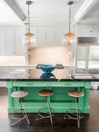 A modern kitchen with a central island painted in a bright green color, topped with a black marble countertop. Three wooden bar stools with metal legs are placed in front of the island. Above the island hang two spherical pendant lights with visible filament bulbs. A decorative blue bowl with intricate patterns sits on the countertop, and a stack of books is placed next to it. The kitchen has sleek white cabinetry, stainless steel fridge, and a light-colored backsplash.