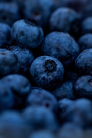 A close-up of ripe blueberries, showcasing their deep blue-purple color and slightly frosted surface. The berries appear plump and fresh, with some displaying small imperfections or natural markings. The focus is centered on the texture and natural details of the fruit.