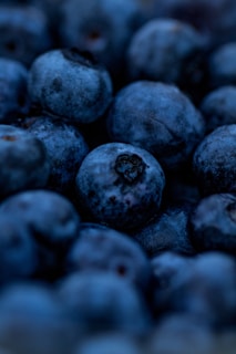 A close-up of ripe blueberries, showcasing their deep blue-purple color and slightly frosted surface. The berries appear plump and fresh, with some displaying small imperfections or natural markings. The focus is centered on the texture and natural details of the fruit.