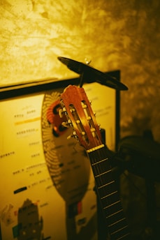 A classical guitar resting against a wall with a mix of soft, warm lighting. The wall features a textured pattern and there is a poster or piece of artwork partially visible behind the guitar.