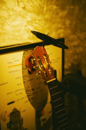 A classical guitar resting against a wall with a mix of soft, warm lighting. The wall features a textured pattern and there is a poster or piece of artwork partially visible behind the guitar.