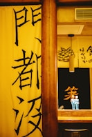 A warm, yellow-themed interior with Japanese characters on banners. There is a wooden pillar in the center and a shelf holding two small, blue figurines. In the background, a drawing of food and additional Japanese text is visible.