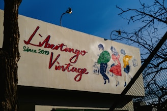 A mural on a building features a colorful illustration of three people walking, each holding a shopping bag. The text 'Libertango Vintage Since 2019' is painted prominently in red and green. The background consists of clear blue sky and some leafless tree branches.