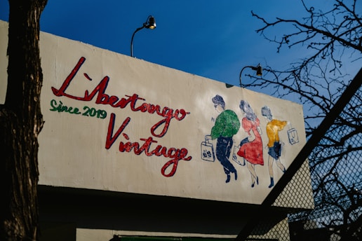 A mural on a building features a colorful illustration of three people walking, each holding a shopping bag. The text 'Libertango Vintage Since 2019' is painted prominently in red and green. The background consists of clear blue sky and some leafless tree branches.