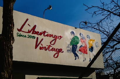 A mural on a building features a colorful illustration of three people walking, each holding a shopping bag. The text 'Libertango Vintage Since 2019' is painted prominently in red and green. The background consists of clear blue sky and some leafless tree branches.
