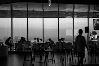A monochrome interior view of a lounge or cafe, featuring large windows overlooking a cityscape. There are several people seated on sofas and chairs, engaged in conversation and working on laptops. Tall bar stools and tables are positioned throughout the room. The lighting appears dim, creating a relaxed atmosphere.