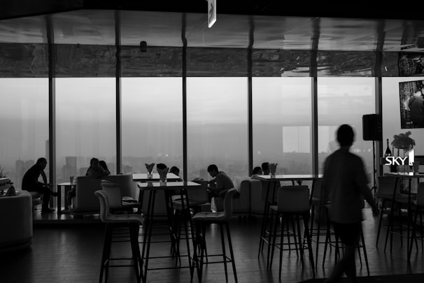 A monochrome interior view of a lounge or cafe, featuring large windows overlooking a cityscape. There are several people seated on sofas and chairs, engaged in conversation and working on laptops. Tall bar stools and tables are positioned throughout the room. The lighting appears dim, creating a relaxed atmosphere.