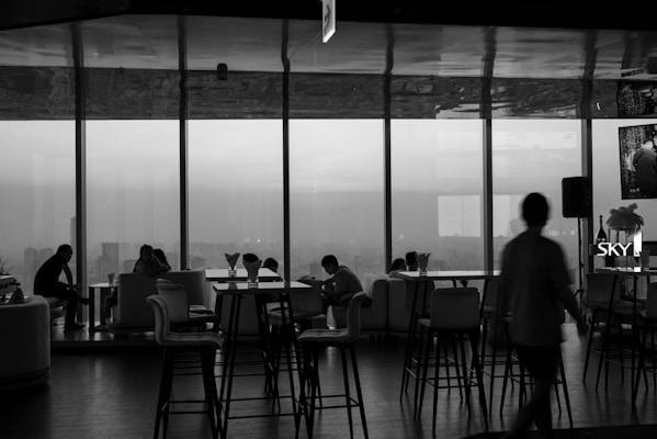 A monochrome interior view of a lounge or cafe, featuring large windows overlooking a cityscape. There are several people seated on sofas and chairs, engaged in conversation and working on laptops. Tall bar stools and tables are positioned throughout the room. The lighting appears dim, creating a relaxed atmosphere.