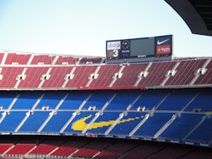 A large football stadium with multiple tiers of red and blue seating. The yellow markings form a logo on the seats. The stadium is mostly empty, with a large electronic scoreboard displaying advertisements and scores.