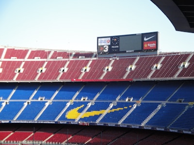 A large football stadium with multiple tiers of red and blue seating. The yellow markings form a logo on the seats. The stadium is mostly empty, with a large electronic scoreboard displaying advertisements and scores.