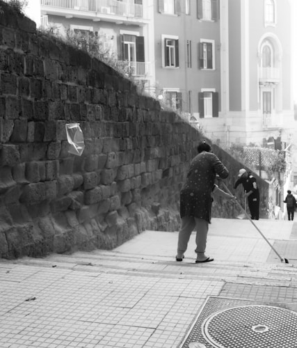 A person is seen sweeping a tiled stairway that runs alongside a stone wall in an urban area. The street is bordered by residential buildings with balconies. Another person in the background appears to be engaged in a similar cleaning activity, and a third person is walking down the steps.