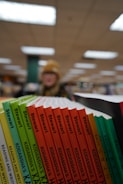 A selection of books with colorful spines is lined up on a shelf. The spines include titles such as 'Stewart' and 'Kodansha's'. The background is blurred, showing a person in a hat with indoor ceiling lights.