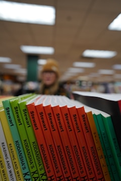 A selection of books with colorful spines is lined up on a shelf. The spines include titles such as 'Stewart' and 'Kodansha's'. The background is blurred, showing a person in a hat with indoor ceiling lights.
