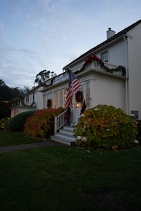 A colonial-style house decorated for the holidays with greenery and red bows. An American flag is prominently displayed near the entrance, flanked by lush, seasonal bushes. The surrounding landscape includes a well-maintained lawn, and trees are visible in the background under a twilight sky.