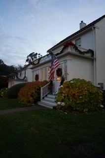 A colonial-style house decorated for the holidays with greenery and red bows. An American flag is prominently displayed near the entrance, flanked by lush, seasonal bushes. The surrounding landscape includes a well-maintained lawn, and trees are visible in the background under a twilight sky.