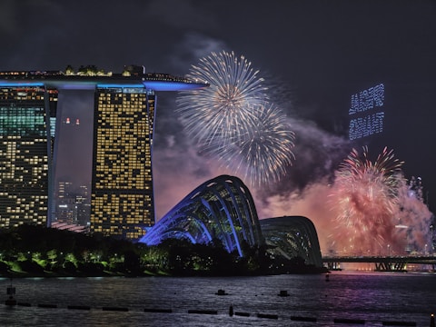 A nighttime cityscape featuring a lit-up, futuristic-looking building complex with a unique rooftop structure, likely the Marina Bay Sands in Singapore. In the foreground, there are arched buildings illuminated in blue. The sky is filled with colorful fireworks, creating an energetic and celebratory atmosphere, with smoke from the fireworks drifting across the scene. The water in the foreground reflects the lights and the fireworks.