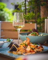 A glass of white wine is positioned on an outdoor dining table surrounded by vibrant greenery. In the foreground, there is a plate of seafood pasta with shrimp and clams, presented with care on a light blue plate. Behind it, a salad with avocado and greens is served in a matching bowl.
