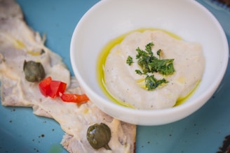 A close-up view of a white bowl containing a creamy dip garnished with green herbs and a drizzle of olive oil. Beside the bowl, there are slices of meat topped with a creamy sauce, capers, and pieces of red bell pepper, all placed on a blue plate.