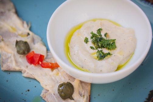 A close-up view of a white bowl containing a creamy dip garnished with green herbs and a drizzle of olive oil. Beside the bowl, there are slices of meat topped with a creamy sauce, capers, and pieces of red bell pepper, all placed on a blue plate.