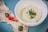 A close-up view of a white bowl containing a creamy dip garnished with green herbs and a drizzle of olive oil. Beside the bowl, there are slices of meat topped with a creamy sauce, capers, and pieces of red bell pepper, all placed on a blue plate.