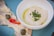 A close-up view of a white bowl containing a creamy dip garnished with green herbs and a drizzle of olive oil. Beside the bowl, there are slices of meat topped with a creamy sauce, capers, and pieces of red bell pepper, all placed on a blue plate.