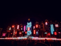 A vibrant cityscape at night featuring an array of brightly lit billboards and neon lights in various colors. The scene is dominated by red, blue, and purple hues, with distinctive text and graphics visible on electronic displays. The atmosphere suggests a lively and futuristic urban environment, bustling with activity and technology.