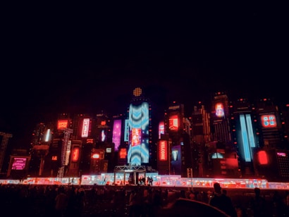 A vibrant cityscape at night featuring an array of brightly lit billboards and neon lights in various colors. The scene is dominated by red, blue, and purple hues, with distinctive text and graphics visible on electronic displays. The atmosphere suggests a lively and futuristic urban environment, bustling with activity and technology.