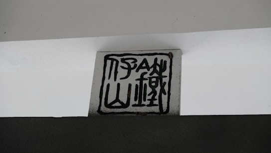 A block with abstract black line patterns on a light gray background is placed against a dark gray wall. The design resembles stylized calligraphy or a symbolic emblem.