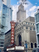 A vintage theatre signboard prominently displays 'Chicago' in bright red letters. Historic architecture with ornate detailing contrasts with modern high-rise buildings. The marquee advertises local events beneath a partly cloudy blue sky.