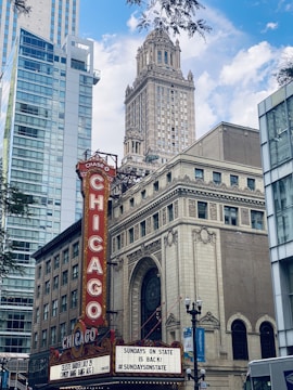 A vintage theatre signboard prominently displays 'Chicago' in bright red letters. Historic architecture with ornate detailing contrasts with modern high-rise buildings. The marquee advertises local events beneath a partly cloudy blue sky.