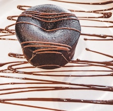A rich, dark chocolate dessert with a smooth surface, elegantly drizzled with chocolate sauce. The dessert sits on a white plate with artistic, flowing lines of chocolate as decoration.
