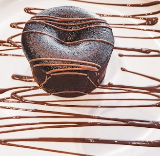 A rich, dark chocolate dessert with a smooth surface, elegantly drizzled with chocolate sauce. The dessert sits on a white plate with artistic, flowing lines of chocolate as decoration.