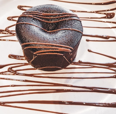 A rich, dark chocolate dessert with a smooth surface, elegantly drizzled with chocolate sauce. The dessert sits on a white plate with artistic, flowing lines of chocolate as decoration.