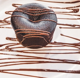 A rich, dark chocolate dessert with a smooth surface, elegantly drizzled with chocolate sauce. The dessert sits on a white plate with artistic, flowing lines of chocolate as decoration.