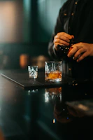 Moody close-up of a bartender’s hands expertly pouring genever into a glass, set against a dark wood bar.