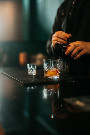 Moody close-up of a bartender’s hands expertly pouring genever into a glass, set against a dark wood bar.