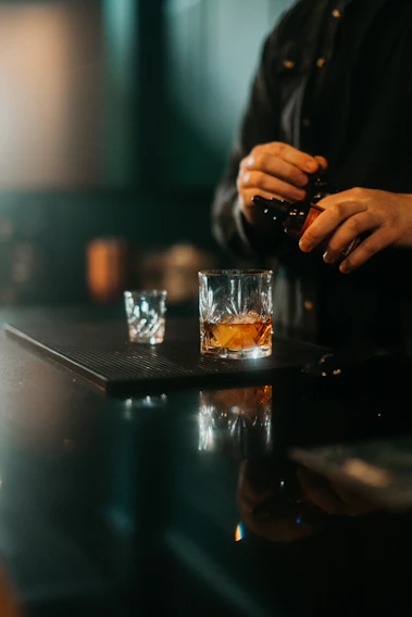 A moody close-up of a bartender’s hands expertly pouring genever into a chilled coupe glass, set against a dark wood bar backdrop.