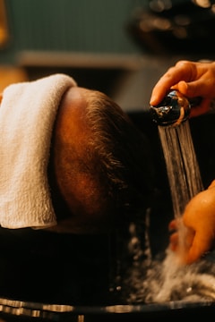 A person is having their head washed in a basin. Water is gently poured from a handheld showerhead, while a towel is wrapped around the forehead to protect the eyes and face. The environment appears to be a salon or barber shop, given the professional care and grooming setup.