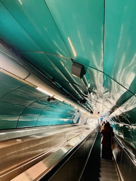 An indoor escalator leads upwards in a tunnel with sleek, reflective, metallic walls and ceilings predominantly in a teal color. There are several lights and fixtures lining the sides of the escalator. A person with long hair and dark clothing is visible towards the lower right on the escalator.