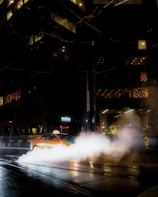 A taxi driving through a city street at night with steam rising from vents in the pavement. The scene is illuminated by the lights from surrounding tall buildings and reflections on the wet road surface.