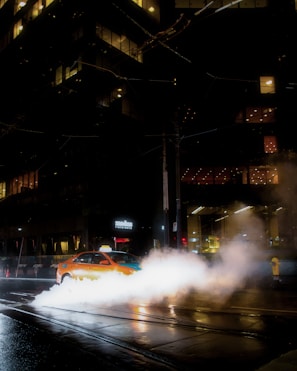 A taxi driving through a city street at night with steam rising from vents in the pavement. The scene is illuminated by the lights from surrounding tall buildings and reflections on the wet road surface.