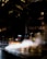A taxi driving through a city street at night with steam rising from vents in the pavement. The scene is illuminated by the lights from surrounding tall buildings and reflections on the wet road surface.
