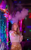 A person standing in a vibrant, colorful setting with neon lights and party decorations. The individual is holding a vaping device, exhaling a large cloud of vapor. The background is decorated with shiny, metallic streamers and balloons, creating a festive atmosphere.