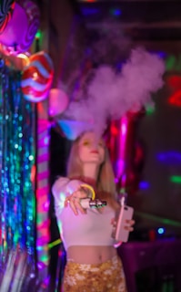 A person standing in a vibrant, colorful setting with neon lights and party decorations. The individual is holding a vaping device, exhaling a large cloud of vapor. The background is decorated with shiny, metallic streamers and balloons, creating a festive atmosphere.
